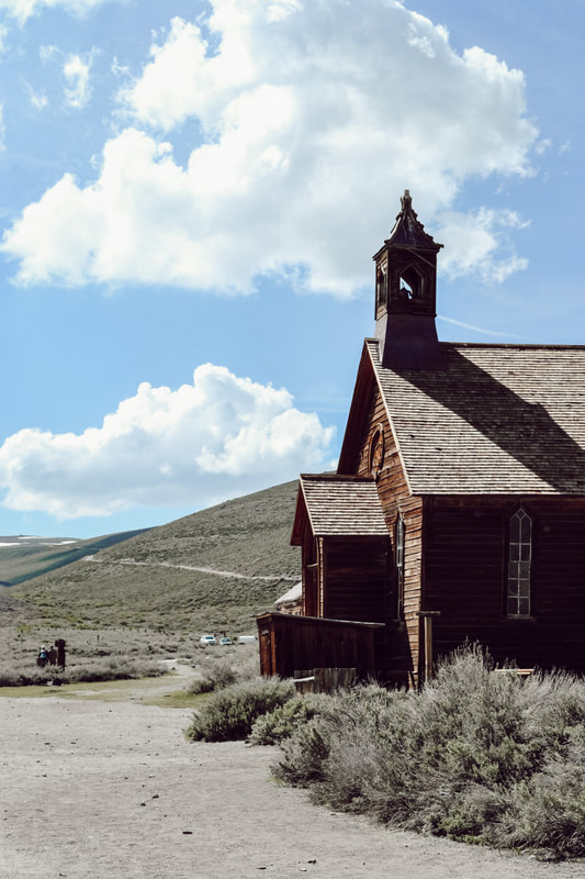 A Picture of the side of a church overlooking a dirt road, with grey-green hills in the background and a cloudy blue sky overhead.