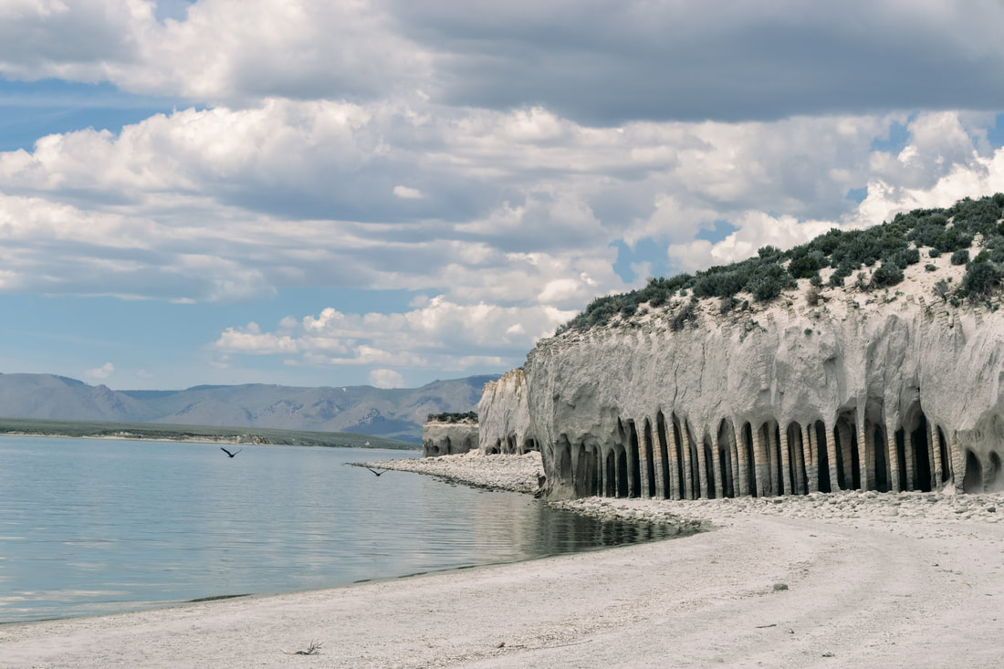 A Picture of Crowley Lake Columns overlooking the lake, with a white sandy beach and the columns to the right, and a pale blue lake reflecting the cloudy sky to the left, with two birds flying above.