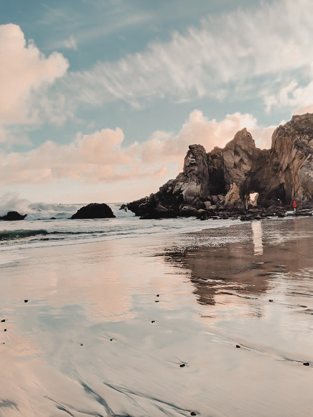 A picture of a keyhole arch at Pfeiffer Beach at sunset, with pink clouds and blue sky reflected in the water and wet sand
